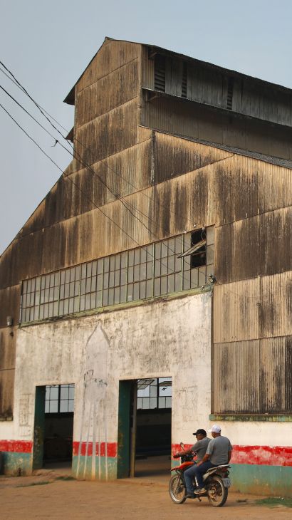 A large warehouse with broken windows sits on a dusty patch of cleared earth.