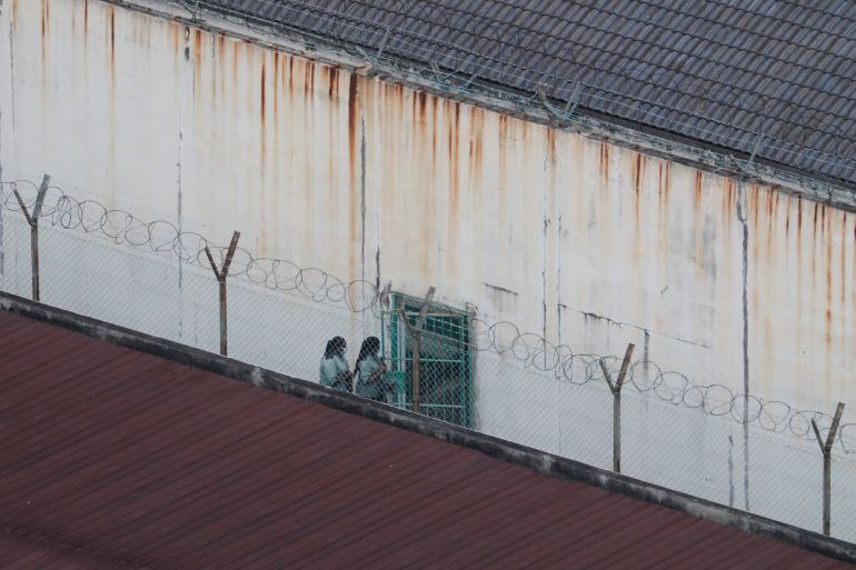 Two people pictured walking inside an immigration detention centre. They are walking against a high wall with high wire fences.