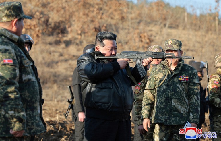 Kim Jong Un holding up a weapon and looking down the barrel. He is wearing a black leather jacket. His hair is slicked back. There are troops in combat fatigues behind him.