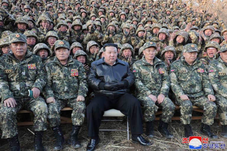 Kim Jong Un sitting with troops. He is wearing black trousers and a black leather coat. The troops are in combat fatigues.