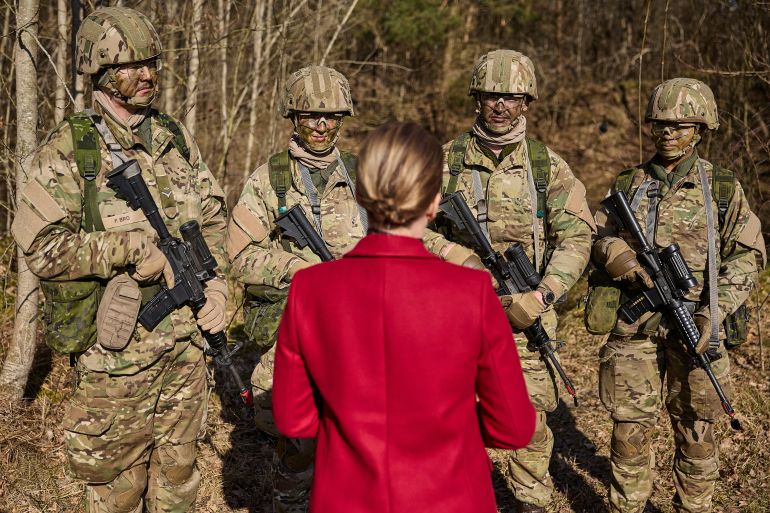 Denmark's Prime Minister Mette Frederiksen speaking to military conscripts. She is wearing a red coat and has her back to the camera. The four conscripts - all male - are in combat uniform and carrying weapons. They are in a forested area.
