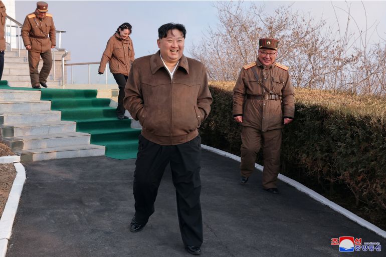 Kim Jong Un at the engine test. He is wearing a brown jacket and black trousers. He has his hands in his pockets. A military officer is nearby and his daughter is walking down the steps behind him.