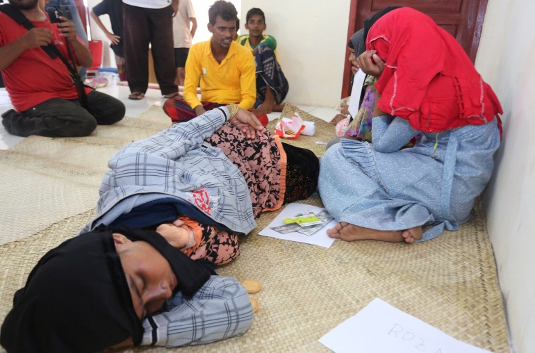 A Rohingya lying on the floor after being rescued from a capsized boat. Some women are seated nearby and look distressed.