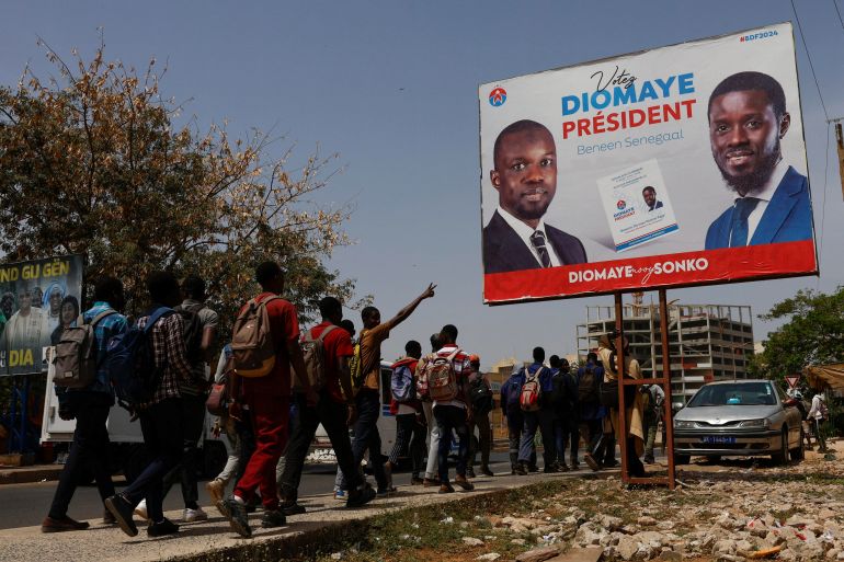 Students walks as they pass an electoral billboard of the Senegalese presidential candidate Bassirou Diomaye Faye, who is backed in the March 24 election by opposition leader Ousmane Sonko, in Dakar, Senegal, March 20, 2024