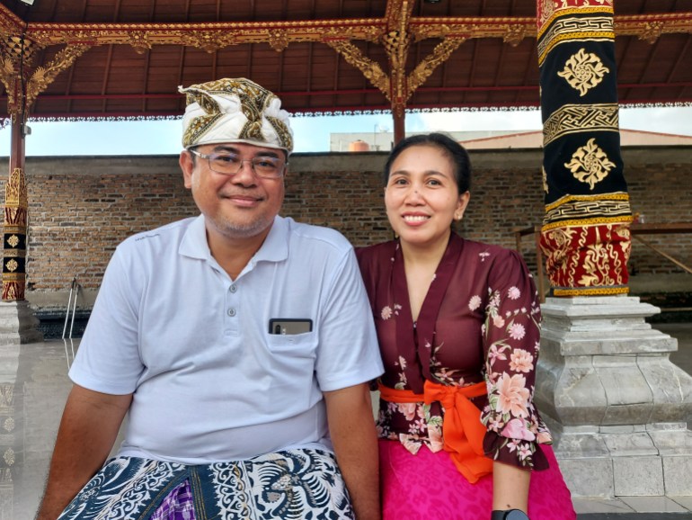 Made Rai and his wife Eni. They are seated on the steps of the temple. Made is wearing a traditional headdress and sarong. Eni is also in a traditional outfit with a sash tied around her waist. They are both smiling.