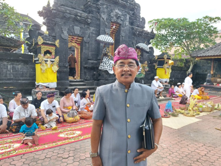  Wayan Dirgayasa standing in the temple. Worshippers are seated on the ground behind him. He is wearing a traditional headdress and jacket., He is smiling and has a bag tucked under this left arm.. 