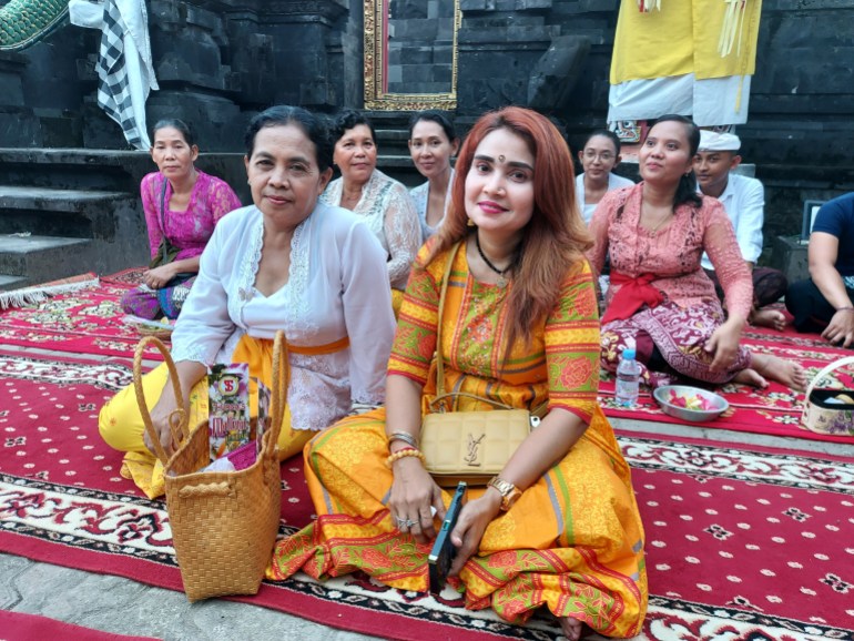 Rajas sitting with a Balinese Hindu woman on a carpet laid out inside the temple courtyard. She is wearing a traditional yellow, red and green floral outfit. The woman next to her is wearing a yellow sarong, with white lace edged top and golden sash. The are smiling.