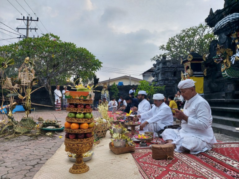 Hindu priests leading the ceremony on the eve of Nyepi. They are wearing white and seated on a carpet. There are offerings in front of them. One is made up of fruit and other objects arranged into a tower.