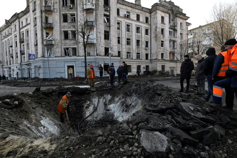 A massive crater on a Kyiv street following a Russian missile attack. There are apartment buildings behind. Workers in high-vis jackets are on site.