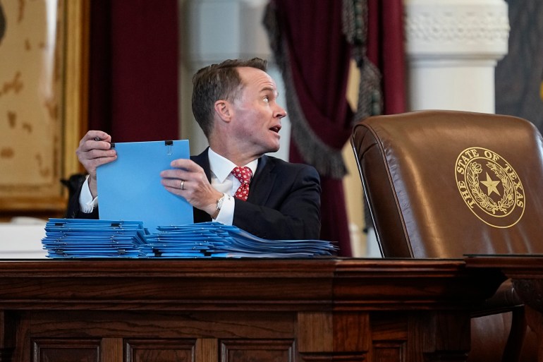Dade Phelan sits behind a large wooden desk in the legislature, with a pile of blue folders before him.