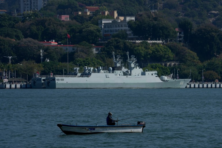 A Chinese guided missile destroyer moored in Xiamen. In the foreground, a man is fishing from a small boat.