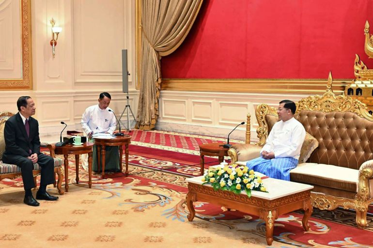 Laos appointed ASEAN special envoy Alounkeo Kittikhoun meets Min Aung Hlaing in Naypyidaw. Min Aung Hlaing is wearing a traditional Burmese outfit and is sitting on a large ornate sofa trimmed in gold. Alounkeo Kittikhoun is sitting at a distance to the general's right in an upright chair. There is a desk with a large flower display in front of Min Aung Hlaing