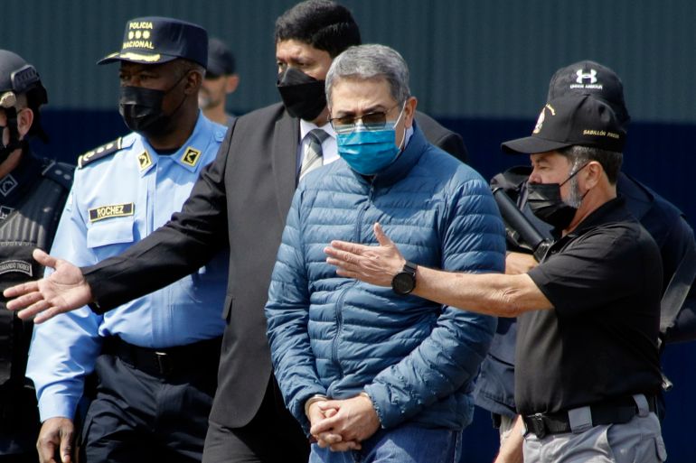 Juan Orlando Hernandez, in a face mask and a blue puffer jacket, is escorted by law enforcement.