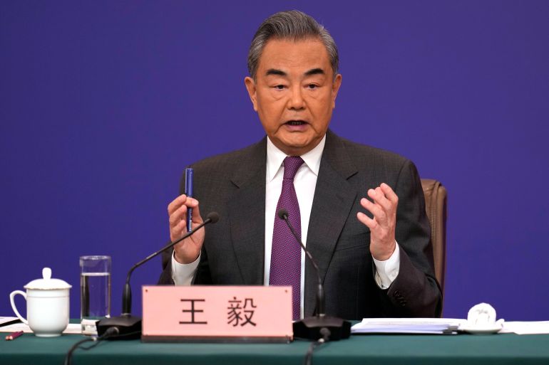 Wang Yi at the press conference in Beijing. He is sitting at a desk against a blue backdrop. He has his hands raised making a point as he speaks.
