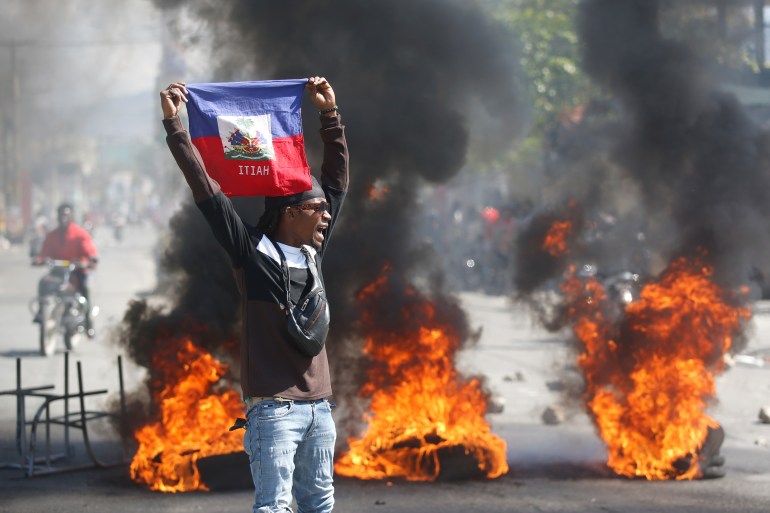A man holding a Haitian flag above his head during unrest in Port-au-Prince. There are fires burning behind him and thick black smoke