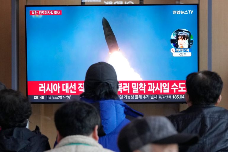 people watch a tv with a rocket launching and red news chyron