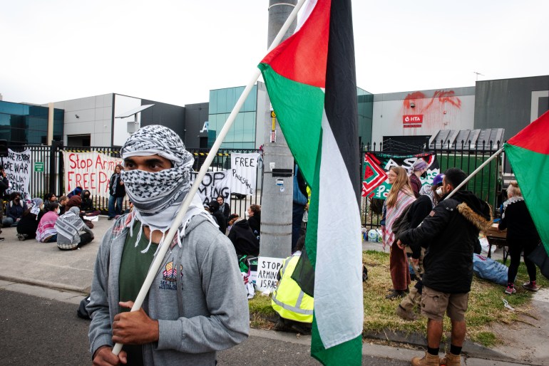 A protester carrying a Palestinian flag at a picket outside an Australian arms company. They have wrapped a Palesinian scarf around their face so only their eyes are visible, Other protesters are behind them. They have placards. Some are sitting on the ground.
