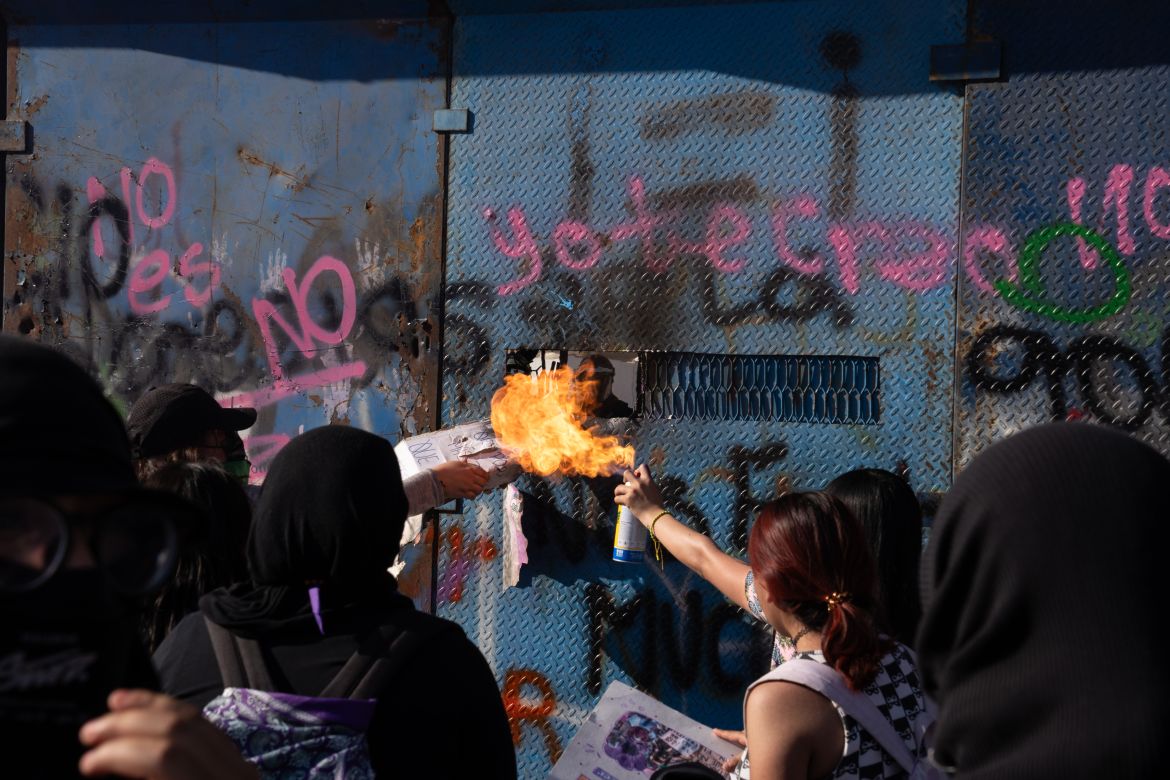 Protesters burn their banners and cards in front of the barriers as police officers in helmets, seen through the window, wait behind the protection of the barriers. Periodically, the police sprayed chemical powder through the gaps to disperse the protesters [Lexie Harrison-Cripps / Al Jazeera]