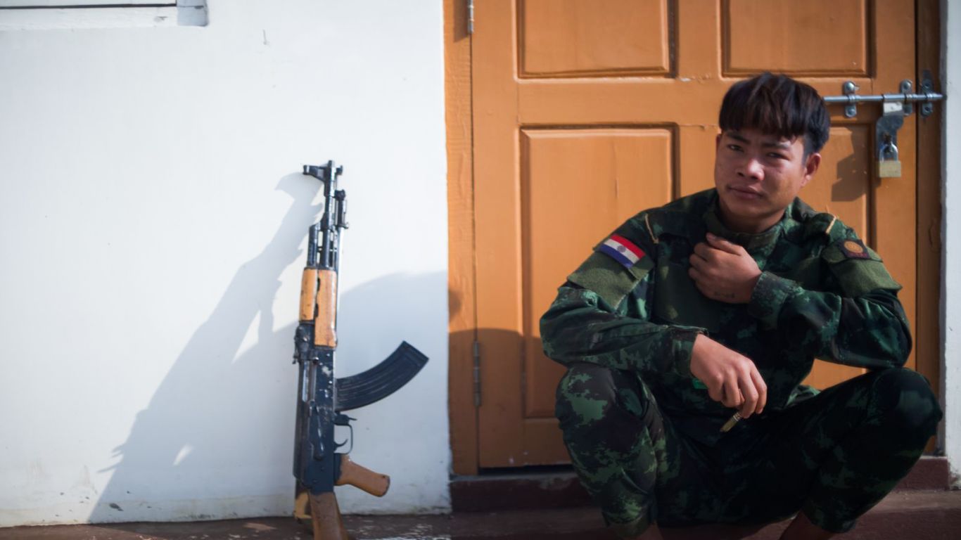 A fighter in Kayah. He is seated on the floor against a door. His weapon is propped against the wall next to him.
