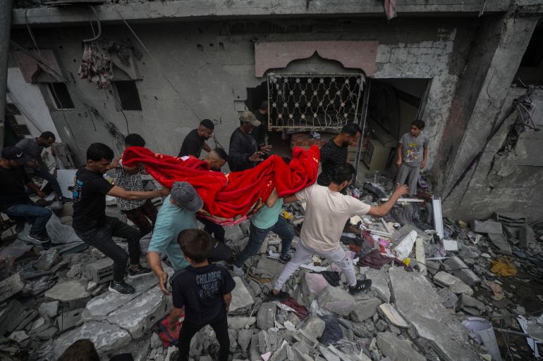 people carry a body wrapped in a red blanket from a destroyed building
