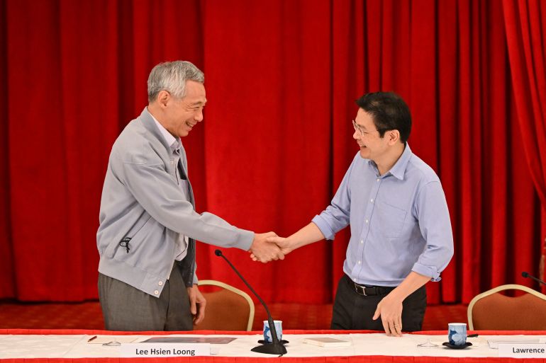 Lee Hsien Loong and Lawrence Wong shaking hands. They are standing behind a desk in front of a red curtain.