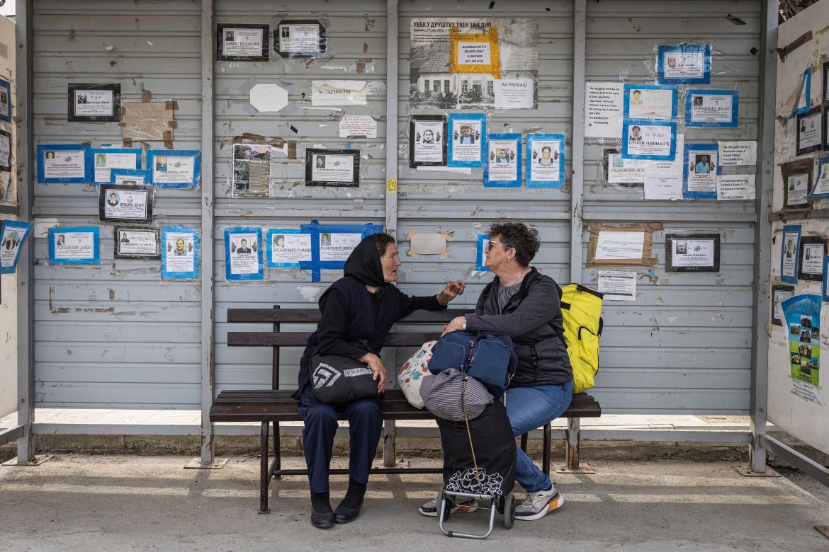 Two women wait at a bus stop, where obituaries are taped up behind them, in the village of Krivelj, Serbia, April 3
