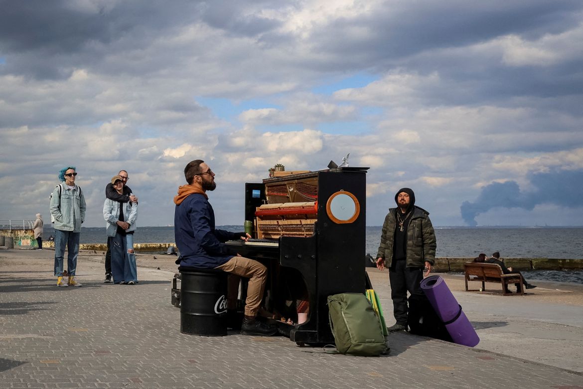 Street musician from Kyiv, Ihor Yanchuk, conducts an impromptu piano concert on the beach to support local residents as smoke rises over the port of Pivdennyi after Russian missiles struck, amid Russia's attack on Ukraine, in Odesa, April 19