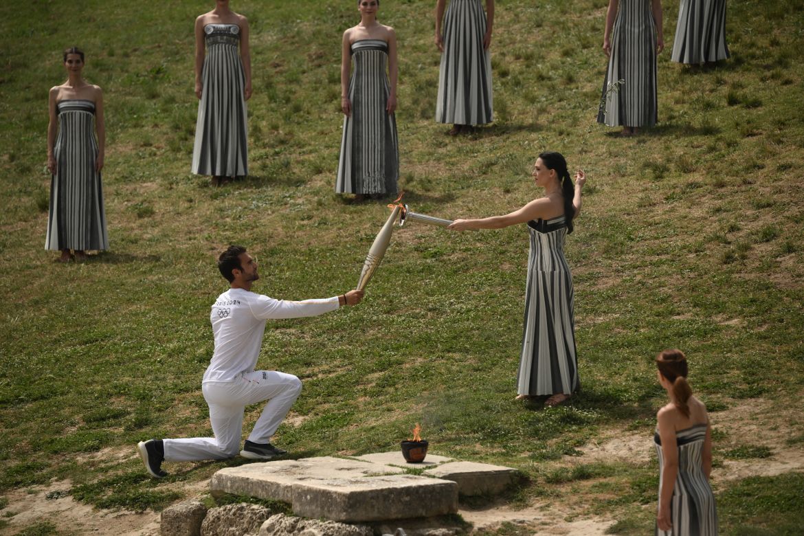 First torch bearer, rowing Olympic gold medalist on 2020, Stefanos Ntouskos (L) receives the flame from Greek actress Mary Mina, playing the role of the High Priestess, as he starts his run with the Olympic torch following the flame lighting ceremony for the Paris 2024 Olympics Games at the Ancient Olympia archeological site, birthplace of the ancient Olympics in southern Greece, on April 16