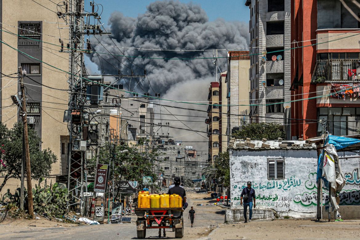 A cloud of smoke erupts down the road from an explosion as a man drives an animal-drawn cart loaded with jerrycans in Nuseirat in the central Gaza Strip on April 17