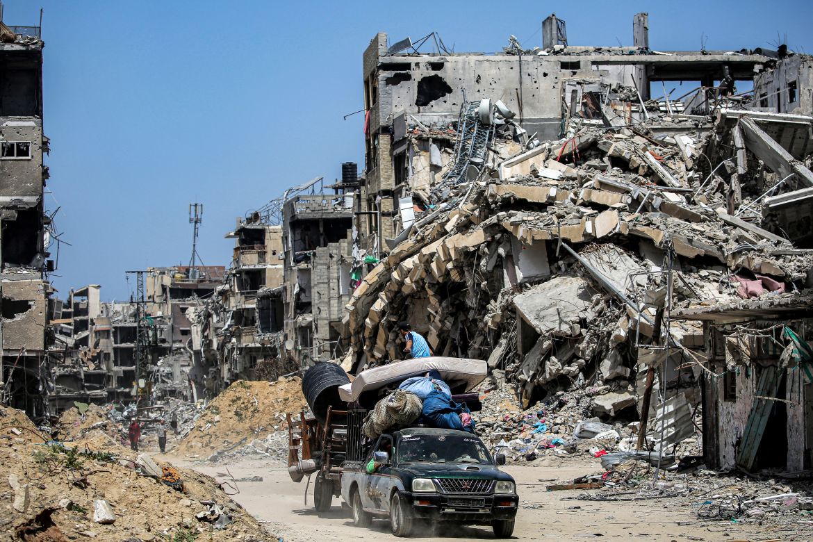 A pickup truck pulling a cart loaded with mattresses and furniture moves past destroyed buildings in Khan Yunis in the southern Gaza Strip