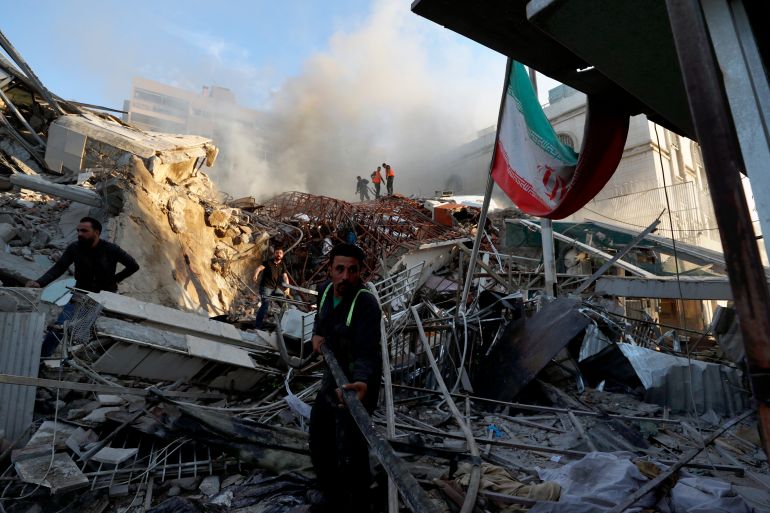 Rescue workers at the scene of the Iran consulate attack in Damascus. The building has been reduced to rubble. There is an Iranian flag to the right.