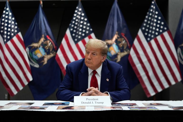 Donald Trump sits in front of a row of US flags, his hands folded. A card indicating his name sits in front of him on the table.