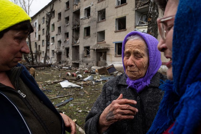 An elderly woman crying after her house was badly damaged in a Russian attack. She has a purple scarf tied around her head and is speaking to two neighbours. There is a bombed out apartment block behind them.