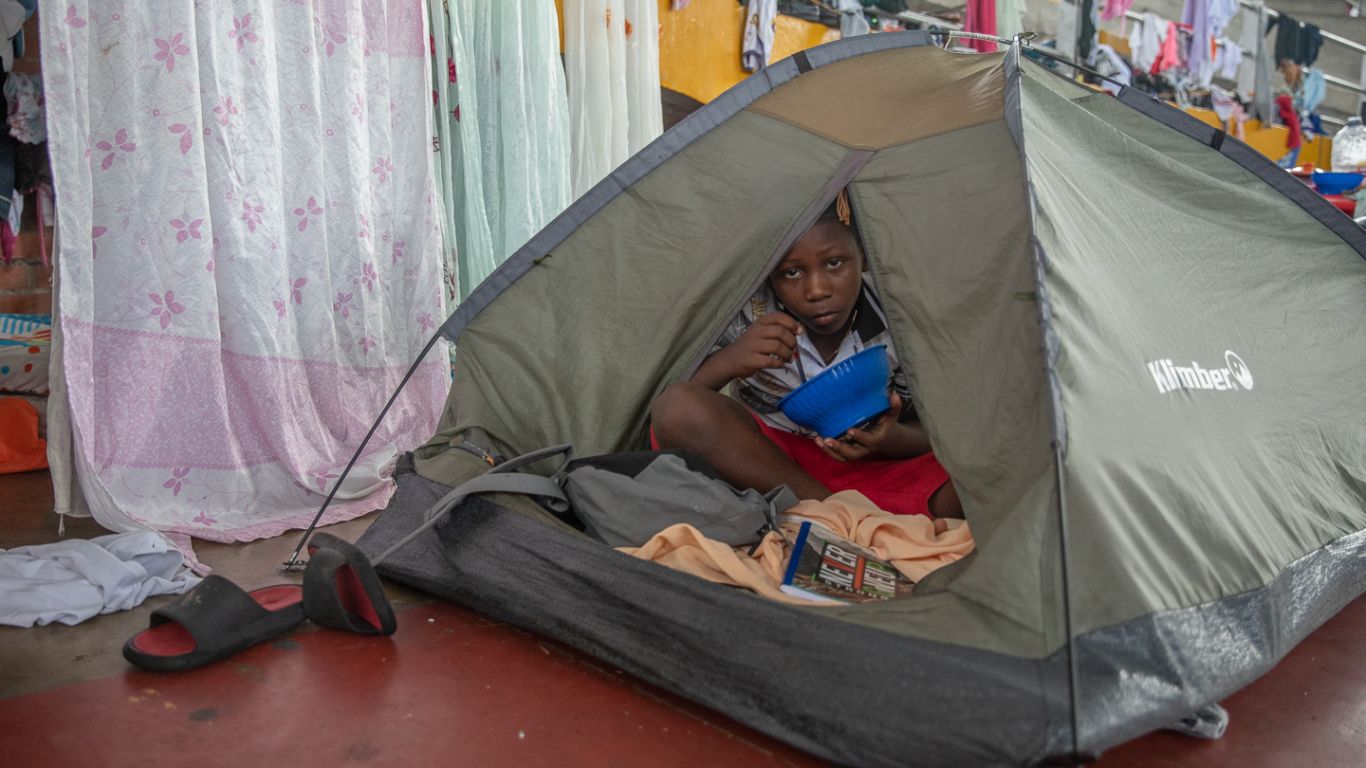A child eating from a bowl peers out from a tent.
