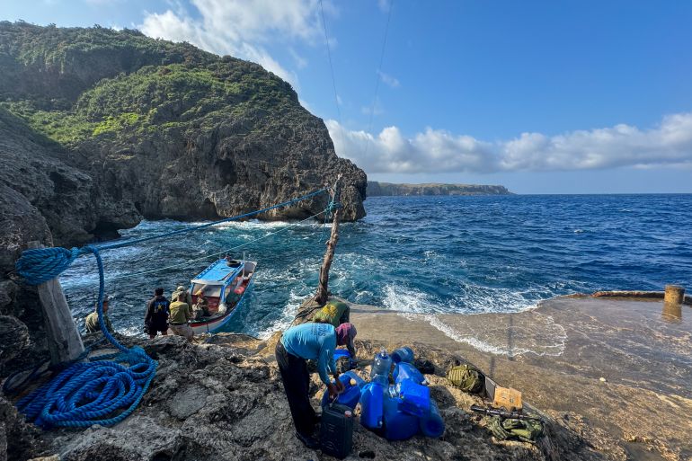 A view of the port in Itbayat. There are jagged cliffs and rocks. A small boat is tied to the shore with ropes, People are waiting to board. The sea looks choppy. The island spreads out behind.