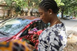 A woman looks at her phone on a pavement while a female e-hailing driver waits in a car in Abuja, Nigeria [File: Afolabi Sotunde/Reuters]