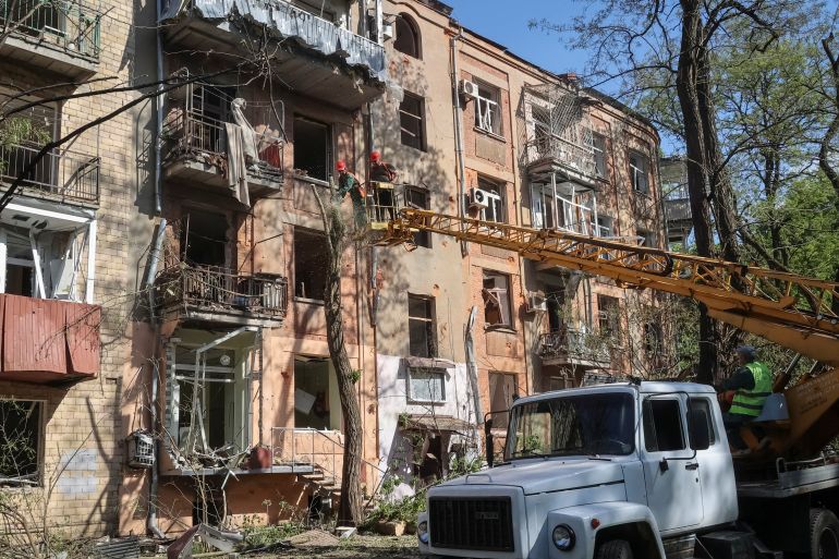 Emergency workers at an apartment building in Kharkiv that was hit in a Russian attack. The building is four storeys high and painted a peachy pink. The windows have been blown out. The workers are on a crane attached to a white truck parked in front.