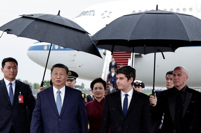 Chinese President Xi Jinping walking beneath an umbrella at Orly airport. He is walking alongside Prime Minister Gabriel Attal. Xi's wife Peng Liyuan in behind him. Their plane is in the background.