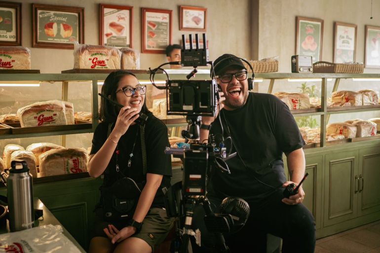 Joko Anwar on set. He is seated next to the camera and has headphones on. He seems to be laughing. A woman is seated on the other side of the camera. They set appears to be a bakery with glass display cases containing bread behind them.