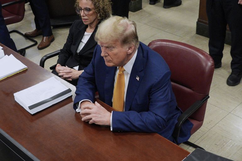 Donald Trump, wearing a blue suit and yellow tie, sits with hands folded at the defence table in the Manhattan criminal court.
