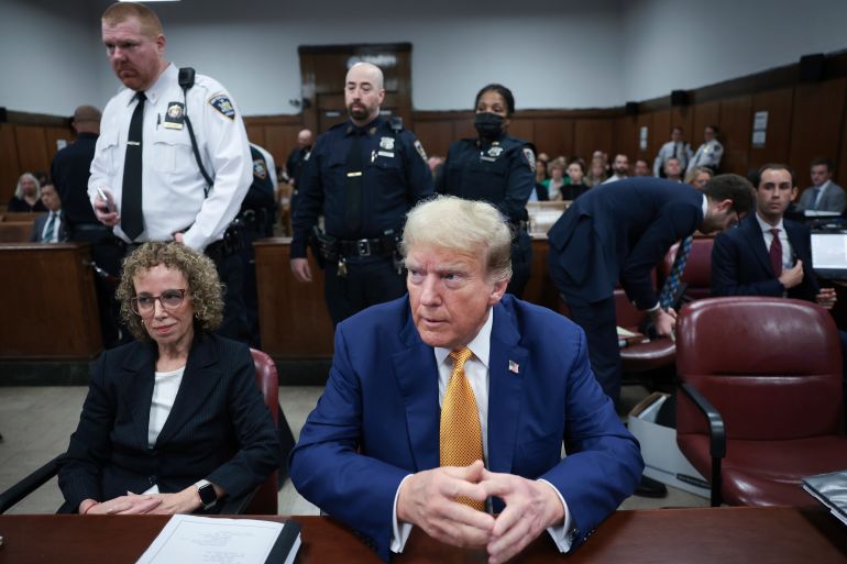 Donald Trump in a New York court. He is sitting at a desk next to his lawyer. He is wearing a blue suit and orange tie. Police and court officials are behind him,