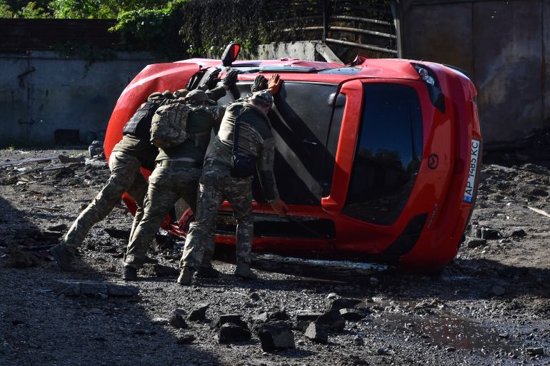 Three Ukrainian soldiers attempting to turn a car the right way up after it was overturned in a Russian missile attack. The car is red and on its side. There is debris around the scene.