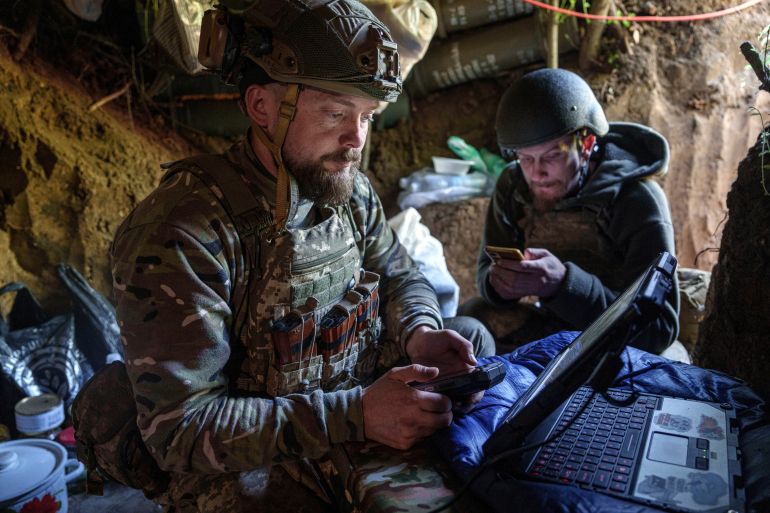 A Ukrainian soldier bear the front line sends drone coordinates. He is seated in a trench. Another soldier is nearby.